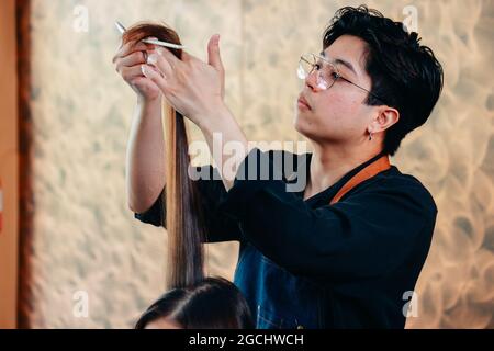 Jeune homme coiffeur asiatique se concentrant sur le travail pendant la coupe et tondre les cheveux longs et magnifiques de la cliente dans un style moderne salon Banque D'Images