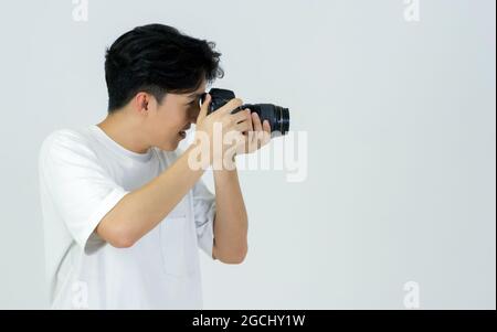 Le jeune photographe asiatique en t-shirt blanc regarde dans le viseur tout en prenant des photos avec l'appareil photo. Ambiance de travail dans le studio photo. Banque D'Images