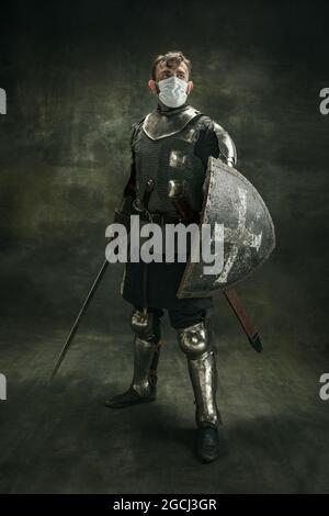 Un homme barbu brutal, un guerrier medeival ou un chevalier dans un masque médical avec bouclier et épée sur fond sombre. Banque D'Images
