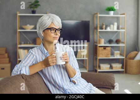 Bonne femme d'âge mûr se détendant sur un canapé, en dégustant une bonne tasse de thé, en regardant loin et en pensant Banque D'Images