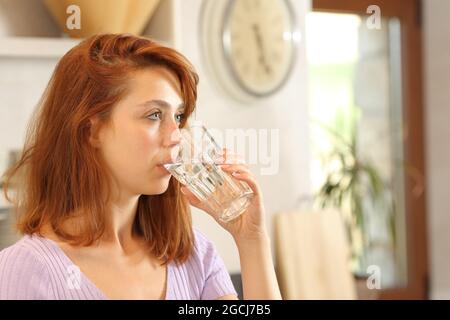 Femme sérieuse qui boit de l'eau du robinet en regardant loin dans la cuisine Banque D'Images