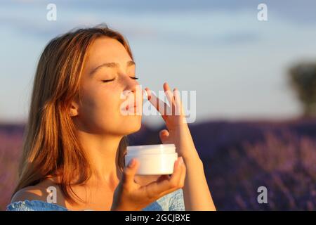 Femme de beauté appliquant la crème hydratante sur la joue dans le champ de lavande au coucher du soleil Banque D'Images