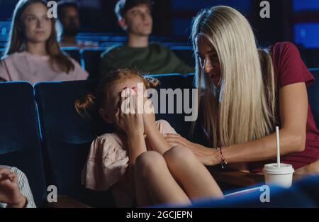 Inquiète petite fille couvrant les yeux dans le cinéma tout en regardant le film, mère réconfortant elle. Banque D'Images