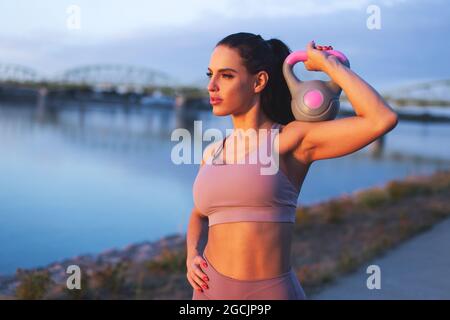 Jeune femme caucasienne tenant la kettlebell sur l'épaule au bord de la rivière, regardant loin, coucher de soleil ou lever du soleil Banque D'Images