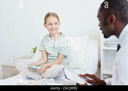 Portrait d'une fille souriante parlant au médecin tout en étant assise sur le lit dans la chambre d'hôpital, espace de copie Banque D'Images