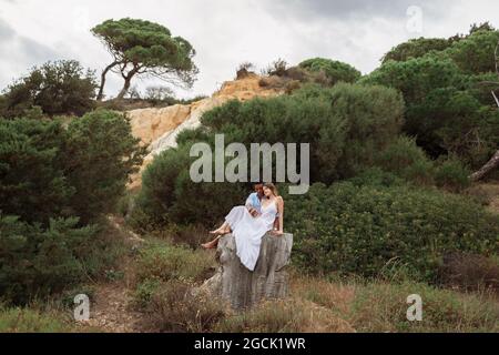 Couple multiracial de jeunes mariés assis sur une souche d'arbre en bois et embrassant le jour du mariage Banque D'Images