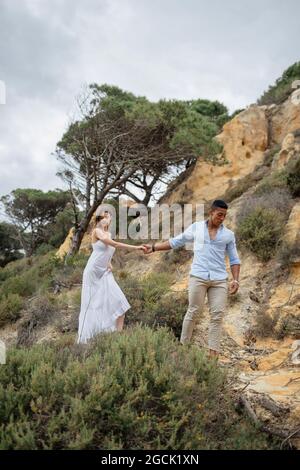 Pont multiracial et marié enchantés tenant les mains et marchant le long de la colline de sable le jour du mariage dans la nature Banque D'Images