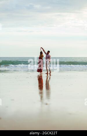 Couple romantique tenant les mains et dansant ensemble sur la plage près de la mer au coucher du soleil en été Banque D'Images
