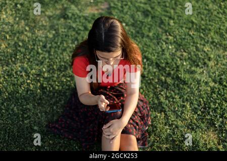 Femme en vêtements d'été assise sur un pré vert dans le parc et surfant sur Internet sur téléphone mobile tout en divertissant le week-end en soirée Banque D'Images