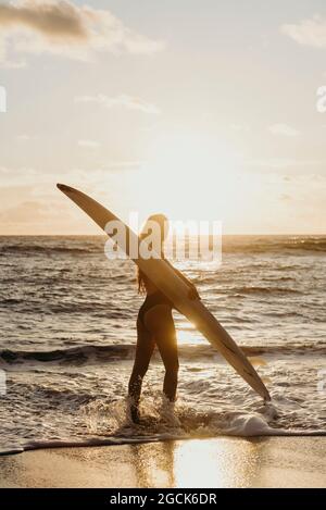 Vue latérale d'une surfeuse féminine méconnue avec promenade en planche de surf dans l'eau de mer au coucher du soleil sur Lanzarote