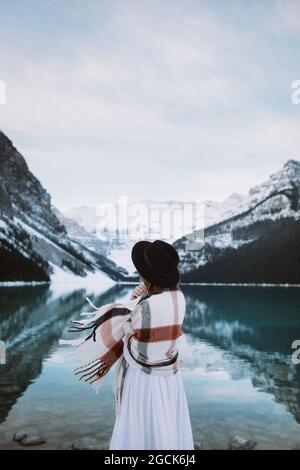 Vue arrière de la femme anonyme en robe blanche et foulard se tenant vers l'eau propre du lac Louise contre la crête de montagne enneigée le jour d'hiver à Alber Banque D'Images