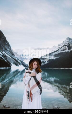 Femme en robe blanche et écharpe debout avec les yeux fermés près de l'eau propre du lac Louise contre la crête de montagne enneigée le jour d'hiver en Alberta, Canad Banque D'Images