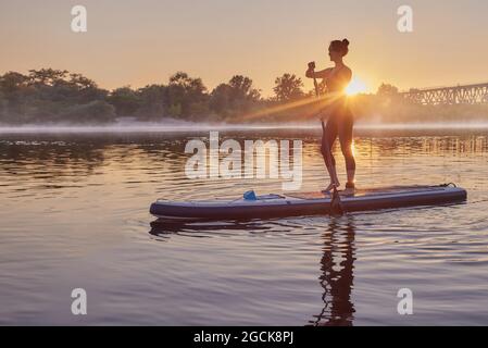 Une femme pagayant sur un panneau SUP sur le fleuve pendant le lever du soleil avec du brouillard au bas. Levez-vous en paddleboard, surfing Banque D'Images