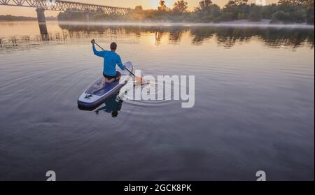 Paddleboard. Homme assis sur un panneau SUP au milieu d'un lac et profitant d'un lever de soleil doré et de brouillard Banque D'Images