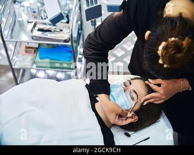 D'en haut crop esthéticienne anonyme avec applicateur couvrant le sourcil de la femme client avec passé pour la lamination pendant le traitement de beauté en salon Banque D'Images