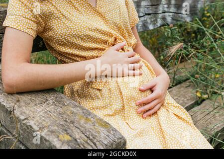 Crop anonyme femme enceinte en robe d'été touchant ventre assis sur le banc dans la campagne en été Banque D'Images