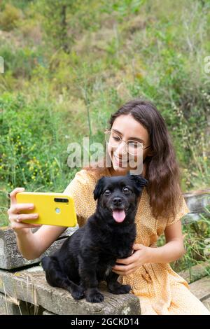 Une jeune femme gaie qui prend sa photo avec un chiot noir moelleux tout en utilisant son smartphone et en étant assise sur un banc en bois à la campagne Banque D'Images