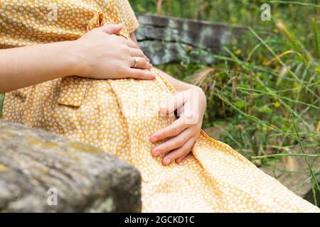 Crop anonyme femme enceinte en robe d'été touchant ventre assis sur le banc dans la campagne en été Banque D'Images