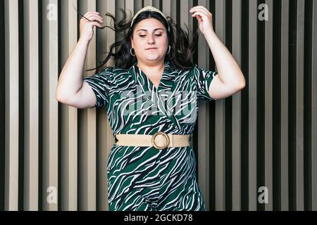 Jeune femme élégante de taille plus, vêtue d'une robe élégante avec décoration géométrique diagonale et ceinture debout, les mains soulevées contre un mur à rayures Banque D'Images