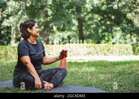 Vue latérale d'une jeune femme fatiguée avec des cheveux foncés dans des vêtements de sport assis sur un tapis sur une pelouse herbacée avec des jambes croisées et de l'eau potable après l'entraînement de yoga en p Banque D'Images