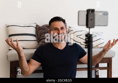 Un homme d'influence enjoué filant sur un téléphone portable sur trépied tout en étant assis au sol à la maison Banque D'Images