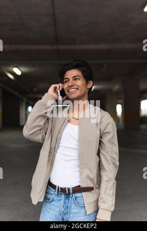 Jeune latin souriant dans des vêtements décontractés parlant au téléphone tout en se tenant dans le parking Banque D'Images
