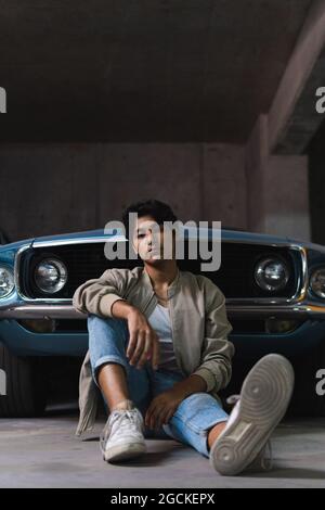 Portrait d'un jeune latino dans des vêtements décontractés regardant avec confiance l'appareil photo tout en s'asseyant sur une voiture d'époque dans le parking Banque D'Images