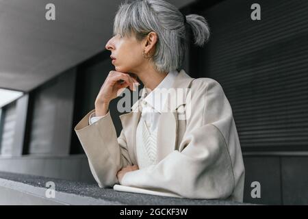 Femme transgenre sérieuse dans un manteau tendance penchée sur une clôture grise tout en se tenant dans la rue en regardant loin Banque D'Images
