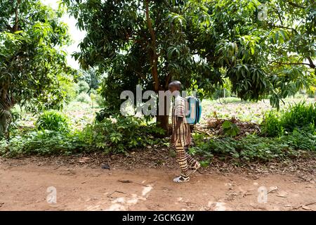 Dans cette image, un écolier africain noir marche seul à l'ombre le long d'une route de terre bordée d'arbres Banque D'Images