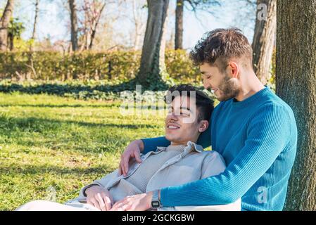 Joyeux couple d'hommes de LBGT embrassant tout en étant assis dans le parc et regardant l'un l'autre Banque D'Images