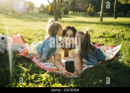 Une jeune femme heureuse et de jolies petites filles allongées sur une couverture et emportant un selfie sur un smartphone tout en s'amusant ensemble sur une prairie verte dans le parc d'été Banque D'Images