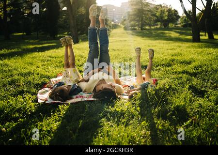 Bonne jeune femme et adorables petites sœurs dans des robes similaires couchés sur une couverture sur l'herbe verte tout en passant la journée d'été ensemble dans le parc et les jambes u Banque D'Images