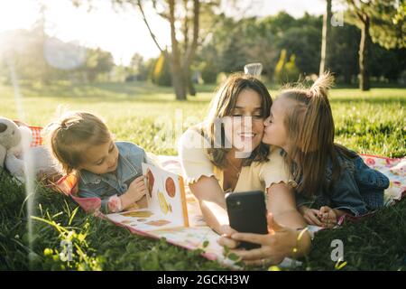 Une jeune femme heureuse et de jolies petites filles allongées sur une couverture et emportant un selfie sur un smartphone tout en s'amusant ensemble sur une prairie verte dans le parc d'été Banque D'Images