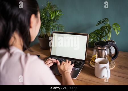 Vue arrière d'une femme asiatique inreconnaissable qui lit des documents sur un ordinateur portable tout en étant assise à table pendant le travail à distance Banque D'Images