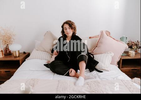 Femme douce en pyjama noir se reposant sur un lit confortable dans la chambre le matin et regardant l'appareil photo Banque D'Images