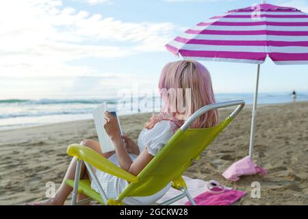Vue arrière d'une femme indépendante anonyme assise dans un fauteuil et travaillant sur une tablette sur une plage de sable près de la mer en été Banque D'Images