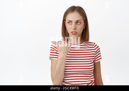 Image de la jeune fille blonde douteuse, frognant et regardant à gauche, pointant vers quelque chose d'incertain, hésitant, étant sceptique au sujet du produit, blanc Banque D'Images