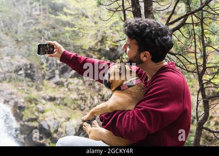 Vue latérale d'un homme joyeux qui se prend en photo avec un Bulldog français sur un smartphone lors d'un voyage dans la forêt montagnarde des Pyrénées Banque D'Images