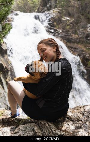 Randonneurs féminins enchantés qui embrassent le Bulldog français près d'une chute d'eau dans les bois tout en appréciant le trekking dans les Pyrénées Banque D'Images