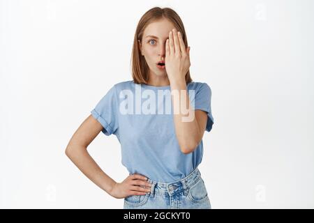 Image de jeune fille blonde cacher la moitié du visage derrière la paume, regardant surpris et étonné d'un oeil, gai et dire wow, en vérifiant la promotion de vente, blanc Banque D'Images