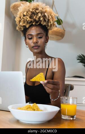 Femme afro-américaine sérieuse utilisant un ordinateur portable et mangeant des chips de maïs avec un verre de jus pour le déjeuner Banque D'Images