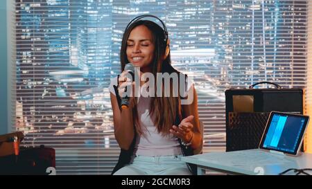 Jolie fille noire dans casque chantant dans le microphone et enregistrer une chanson dans le studio à la maison avec gratte-ciel en arrière-plan. Banque D'Images