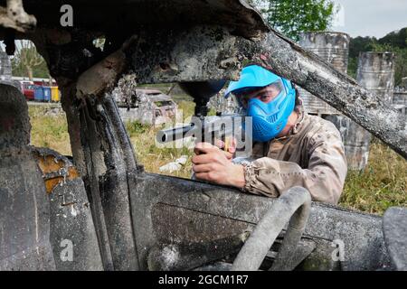 Un joueur masculin dans un casque et avec un fusil jouant du paintball et se cachant derrière une voiture abandonnée de sorbby pendant le match Banque D'Images