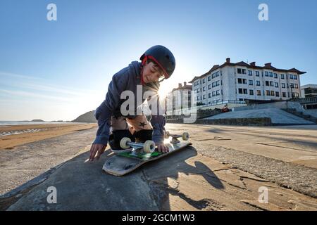 Adolescent gai garçon dans un casque assis avec un skateboard sur la promenade près de la mer en été Banque D'Images