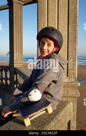 Vue latérale d'un adolescent ravie dans un casque de protection et avec un skateboard assis sur une clôture en pierre près de la mer et regardant la caméra Banque D'Images