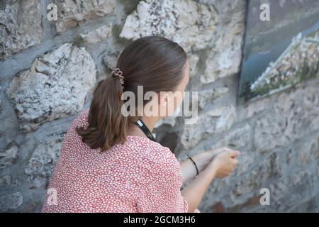 Vue arrière d'une fille touristique dans une robe rouge se reposant à l'ombre à côté d'un vieux mur de pierre lors d'une chaude journée d'été. Banque D'Images