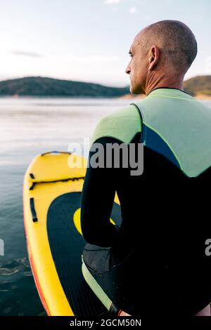 Vue arrière d'un homme adulte en combinaison agenouillée sur un paddle-board sur une surface d'eau calme du lac Banque D'Images