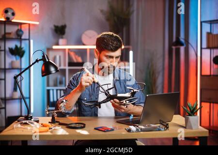 Homme barbu focalisé assis à table et regardant le tutoriel en ligne sur la réparation de drone. Jeune homme regardant sur écran d'ordinateur et de soudure quadcopter. Banque D'Images