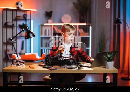 Adolescent de race blanche utilisant un ordinateur portable moderne pour chercher la façon de réparer une voiture cassée. Enfant mignon assis à la table et essayant de réparer le jouet télécommandé à la maison. Banque D'Images