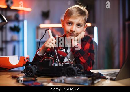 Portrait d'un garçon caucasien dans une tenue décontractée brasant une voiture télécommandée cassée à la maison. Un enfant mignon assis à une table et répare son jouet préféré. Banque D'Images